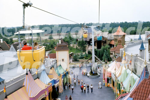 x A 1970s color photo of yellow and blue Skyway buckets traveling over the colorful striped tents of Fantasyland at Walt Disney World in Florida.