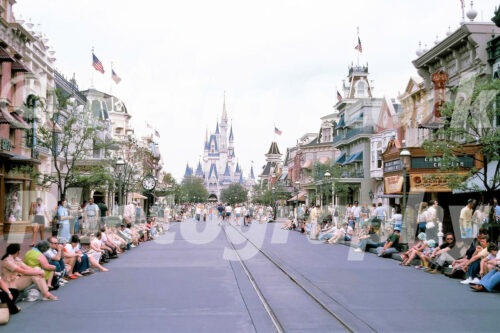 A 1970s color photo of Main Street, U.S.A. at Walt Disney World in Orlando, Florida, featuring Cinderella Castle at the end of the street and guests sitting on curbs waiting for a parade.