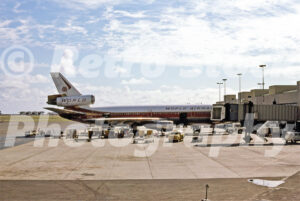 A 1980s color photo of a World Airways McDonnell Douglas DC-10 parked at a jet bridge at Honolulu International Airport in Hawaii with ground service vehicles.