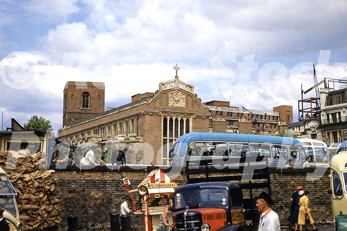 A 1950s colour photo of All Hallows-by-the-Tower church in London, featuring a blue double-decker bus, a red vintage lorry, and a bustling street market in the foreground.