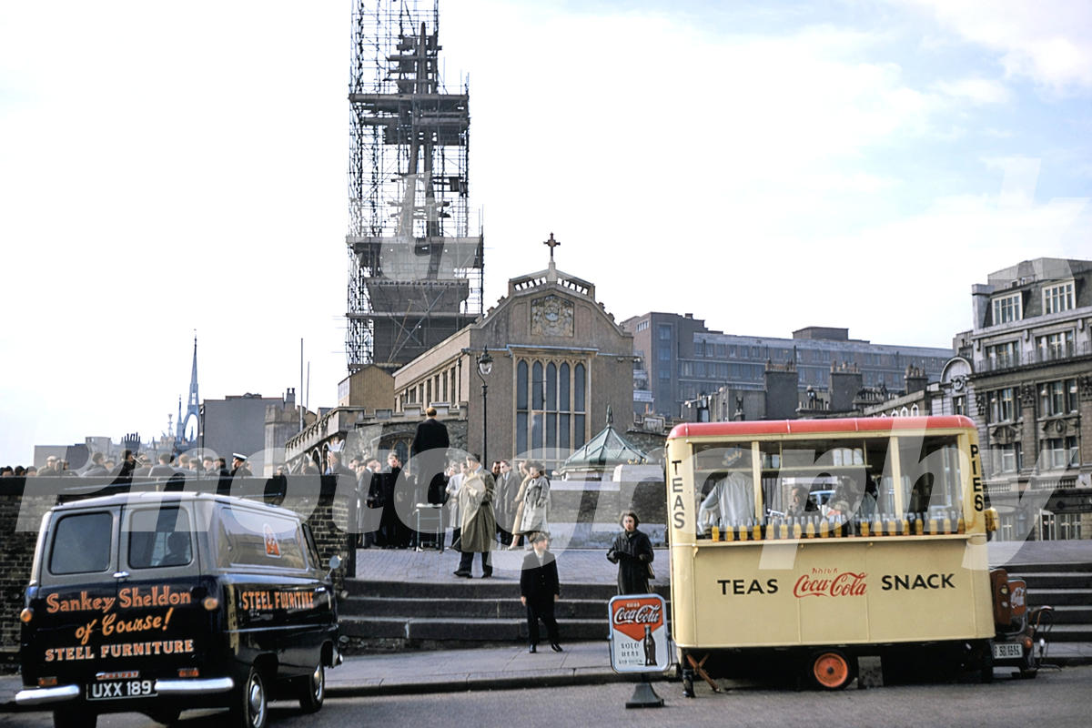 A 1950s colour photo of All Hallows-by-the-Tower in London, showing the church spire surrounded by scaffolding with a vintage tea stall and a Sankey Sheldon delivery van in the foreground.