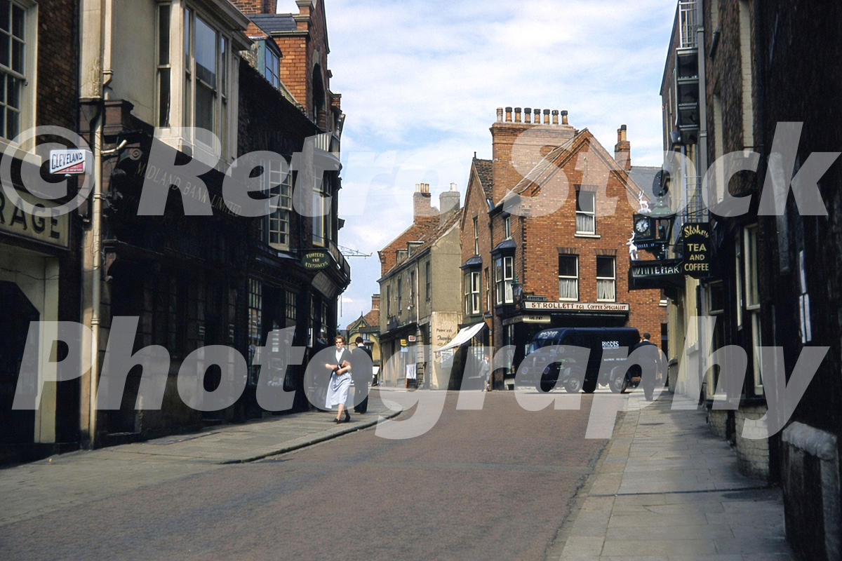 1950s photo of Bailgate in Lincoln, with Midland Bank on the left, shop signs ahead, a van near the junction and pedestrians on the street.