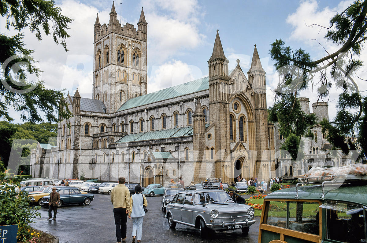 1970s photo of Buckfast Abbey in Devon, with visitors, parked cars including an Austin 1800 Landcrab, and the abbey’s west front seen from the forecourt.