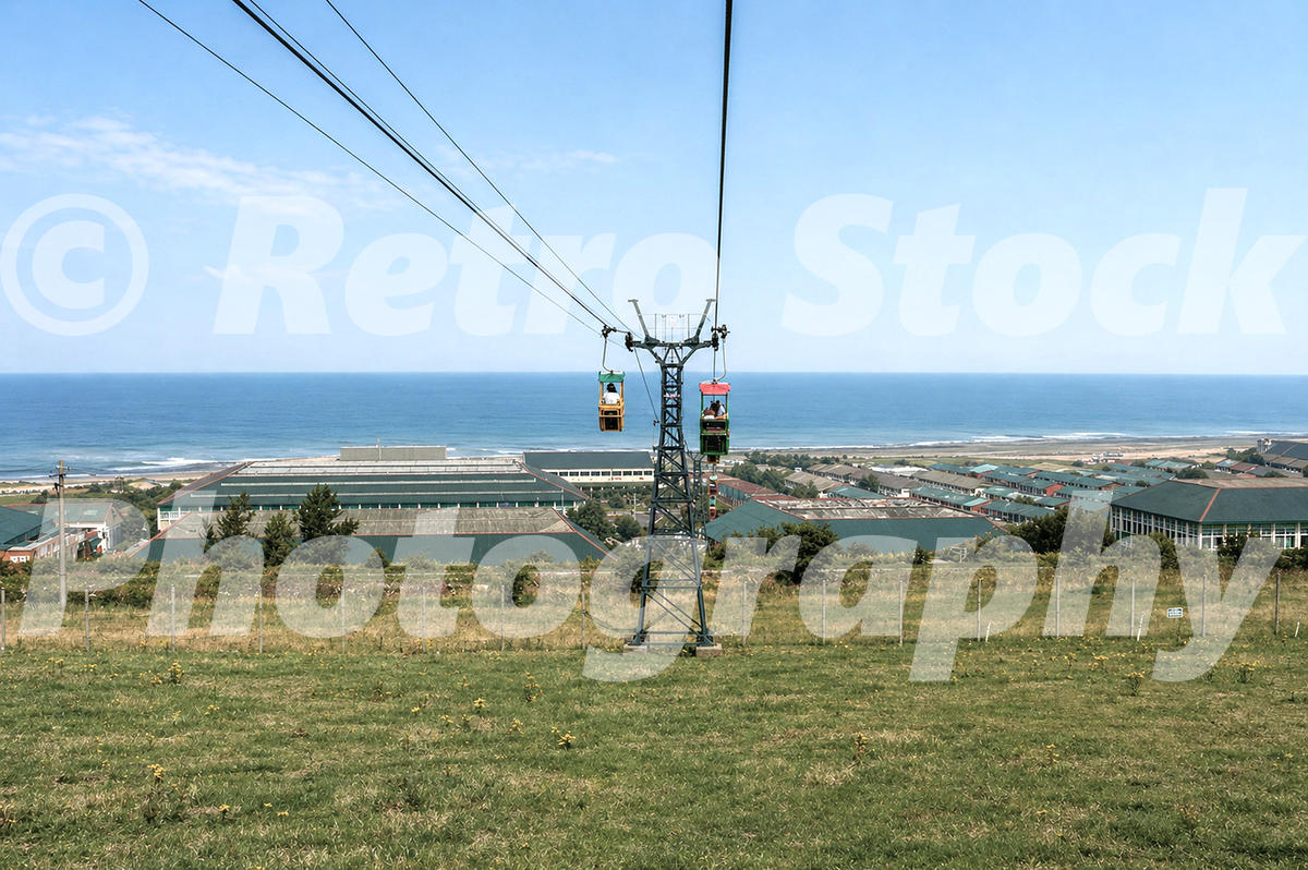 A 1983 colour photograph taken from a chairlift looking down a grassy hillside toward the sprawling Butlins Ayr holiday camp and the sea, with two colorful chairlift cars visible on the cables.