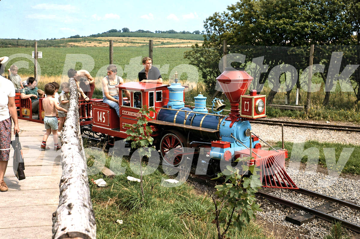 A 1983 colour photograph of a red and blue Chance CP Huntington miniature locomotive (No. 145) stopped at a rustic station platform at Butlins Ayr, with passengers boarding and fields in the background.