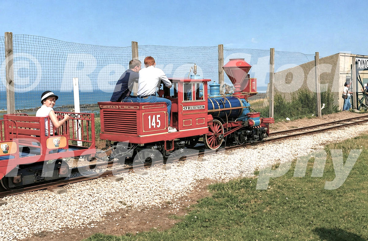 A 1983 colour photograph of a red and blue Chance CP Huntington miniature locomotive (No. 145) carrying passengers along the seaside tracks at Butlins Ayr.