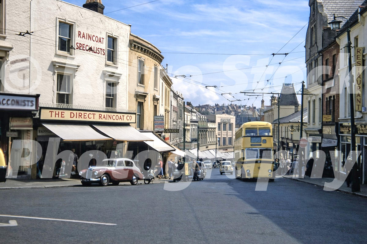 1950s view of Commercial Road in Bournemouth with shops, trolleybus wires, cars, pedestrians and a yellow double-decker trolleybus.