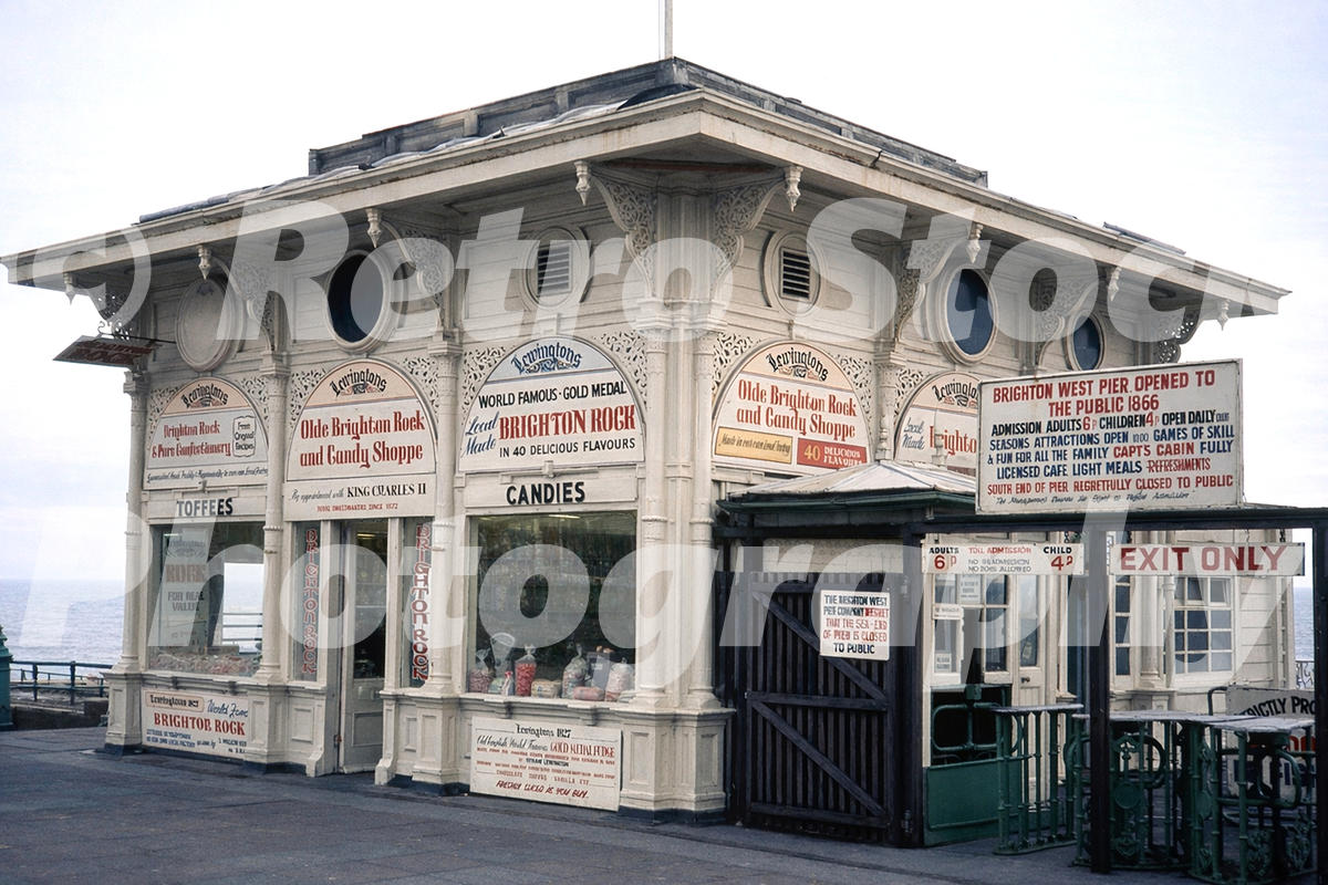 A 1970s photograph of the entrance to the West Pier in Brighton, featuring the ornate white Victorian ticket office and candy shop with numerous signs for Brighton Rock, toffees, and candies.