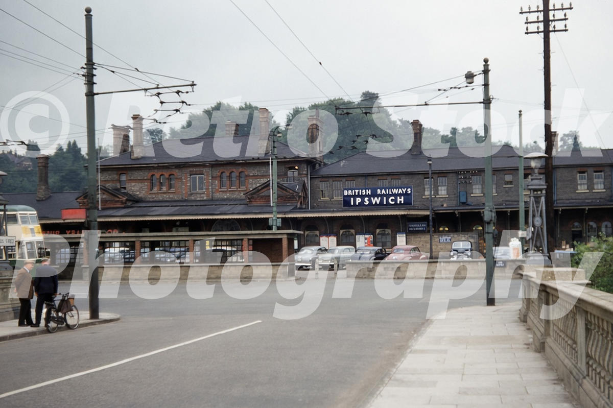 1950s view of Ipswich railway station with British Railways sign, trolleybus wires, parked cars and men beside a bicycle.