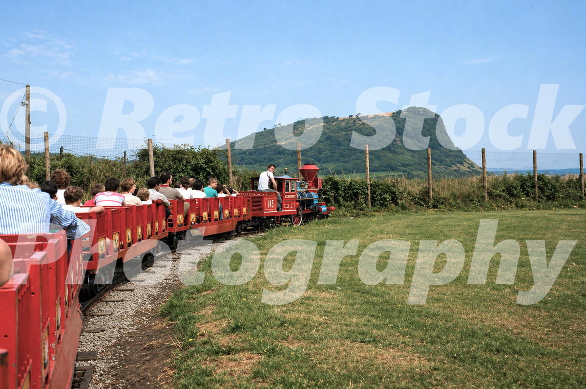 1980s view of the miniature railway at Butlin’s Ayr holiday camp in Scotland, with passengers in red carriages and the Heads of Ayr in the background.
