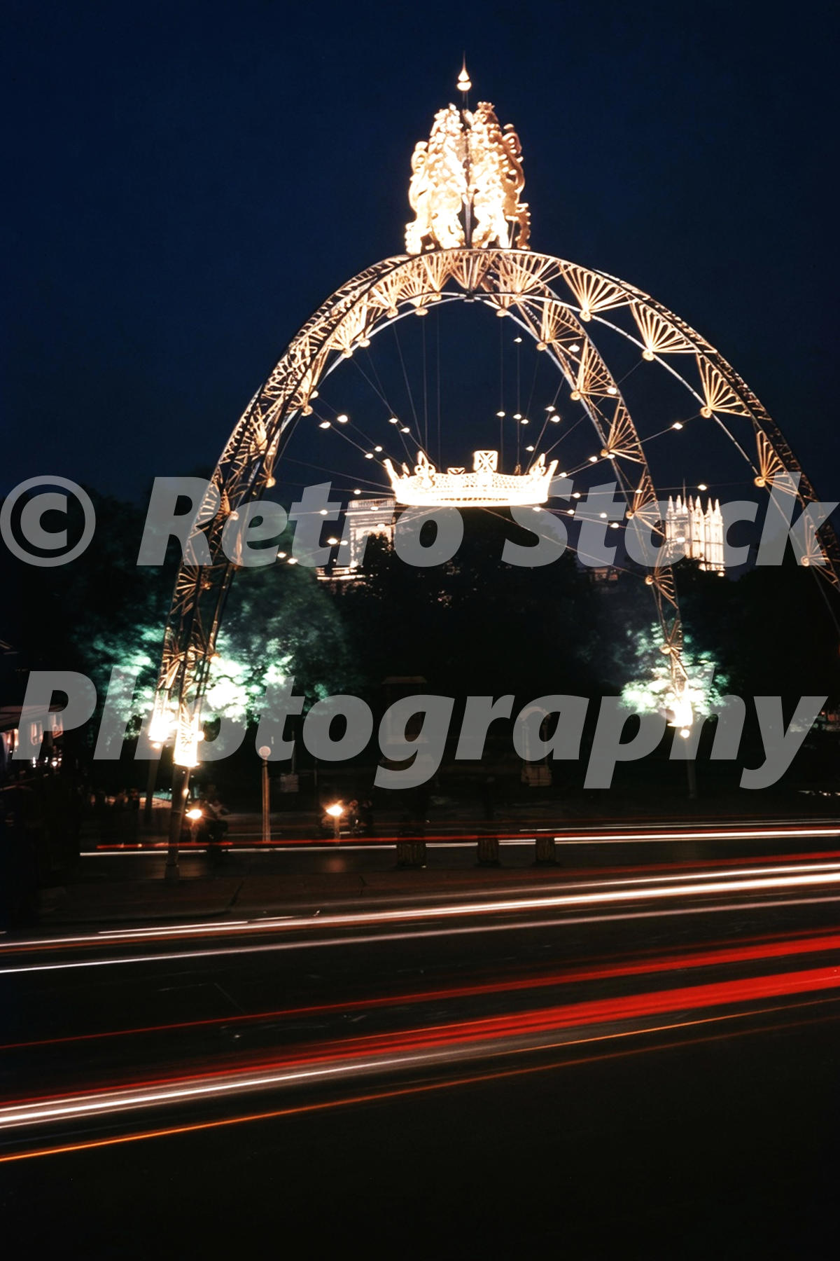 1950s night view of The Mall in London with coronation illuminations, a large lighted arch, crown decoration and traffic light trails.