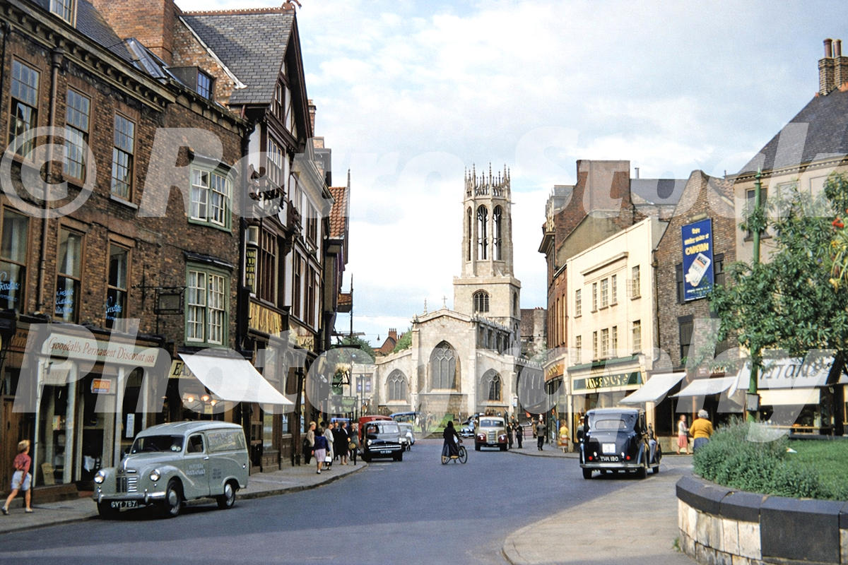 1950s view of Pavement in York, with All Saints Church, shops, pedestrians, vans, cars and a cyclist.