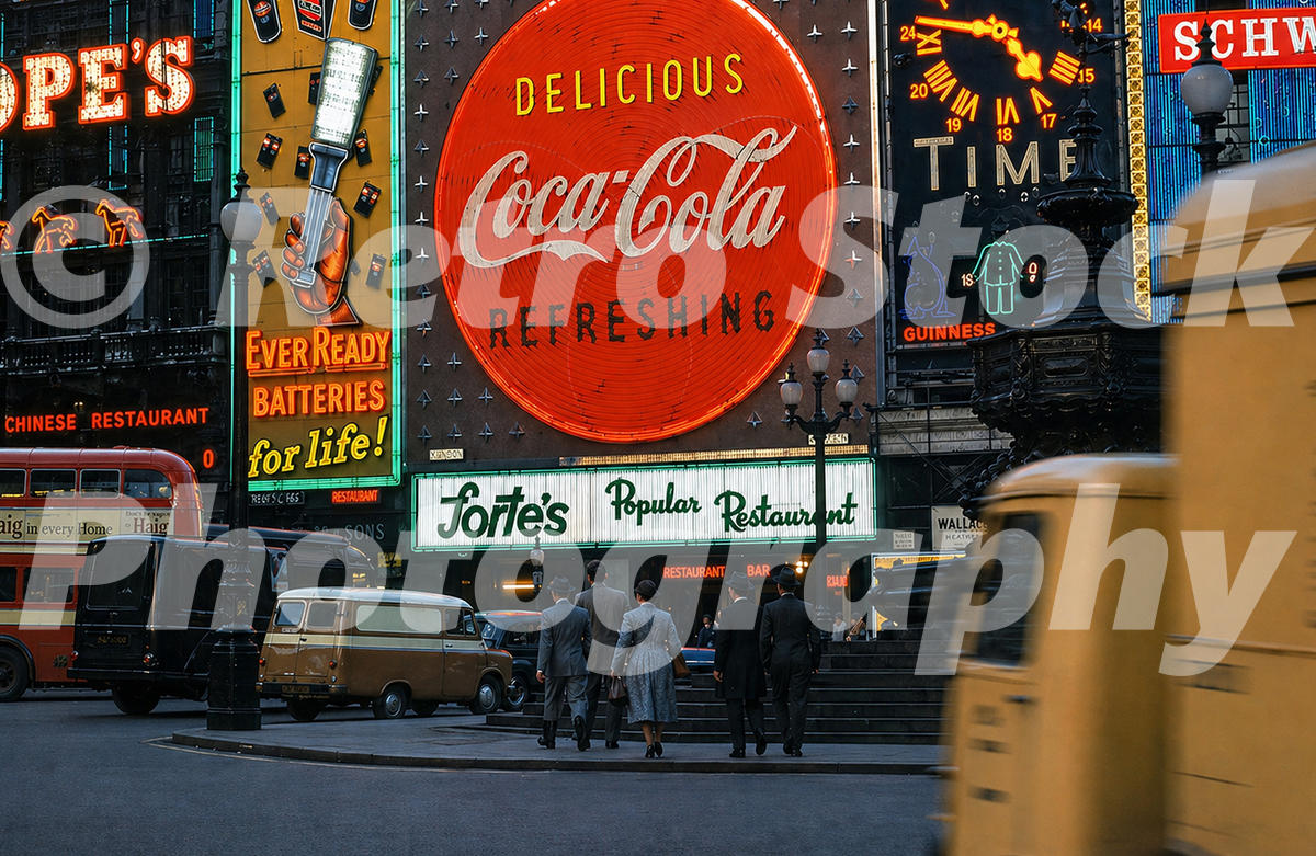 A 1950s colour photo of Piccadilly Circus in London at night, featuring vibrant neon signs for Coca-Cola, Forte's Popular Restaurant, and Ever Ready Batteries.