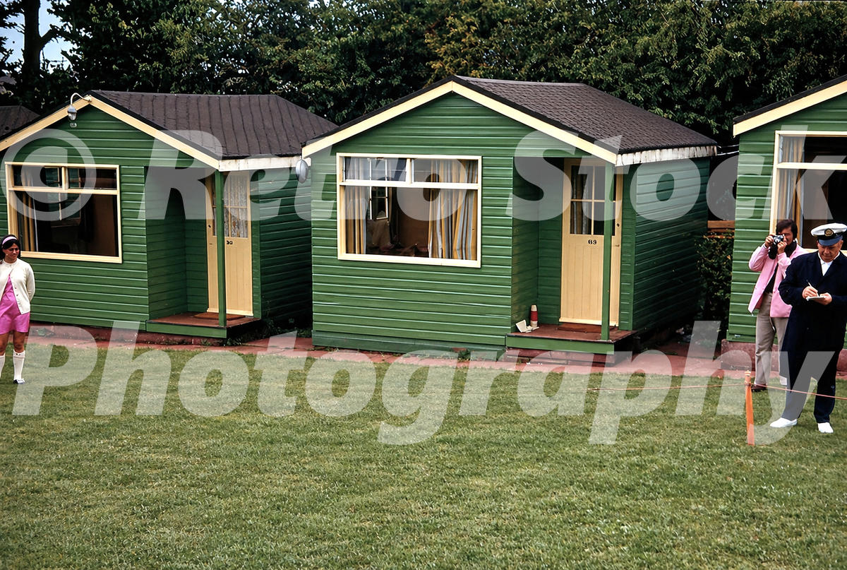 1960s view of green wooden chalets at Pontins South Devon Holiday Camp in Paignton, with numbered doors, striped curtains and people outside.
