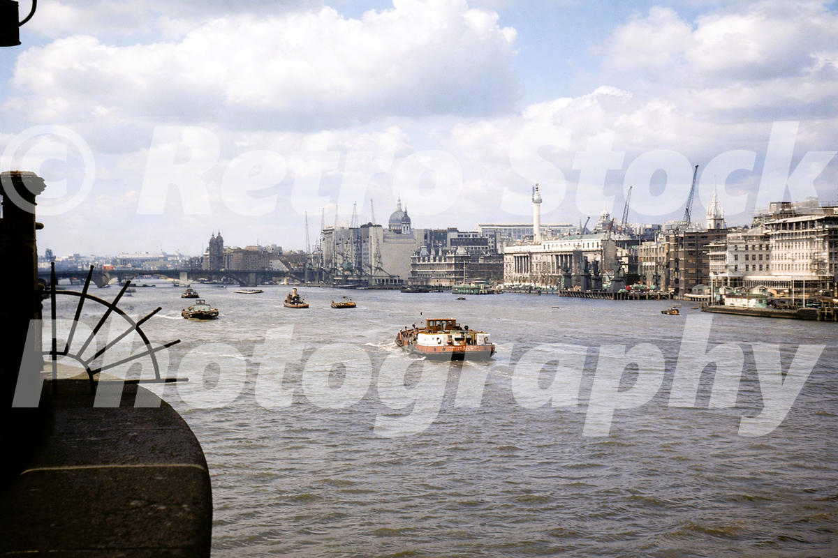 1950s view from Tower Bridge looking along the River Thames into the Pool of London, with boats, cranes, wharves and the City skyline.