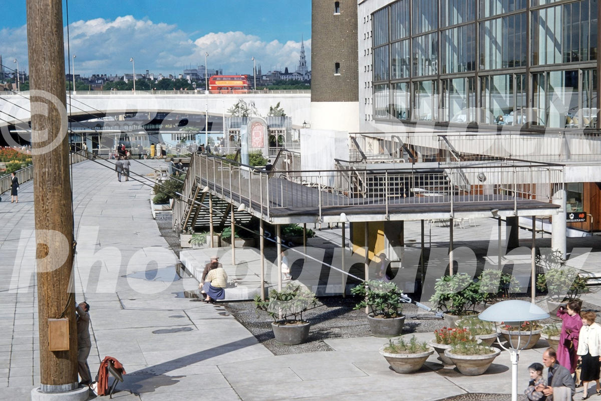 1950s view of Royal Festival Hall and the South Bank in London, with modern terraces, pedestrians, planters and a red bus in the background.