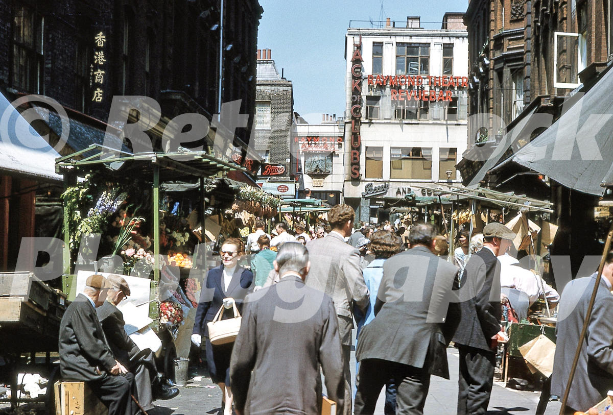 rupert-street-london-1959_55231896498_o 1950s view of Rupert Street market in Soho, London, with crowded stalls and Raymond Revuebar signs in the distance.