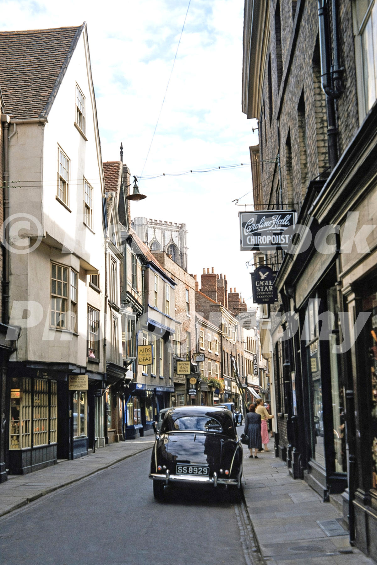 1950s view along Stonegate in York, with York Minster in the distance, a parked black car, shop signs and historic buildings.