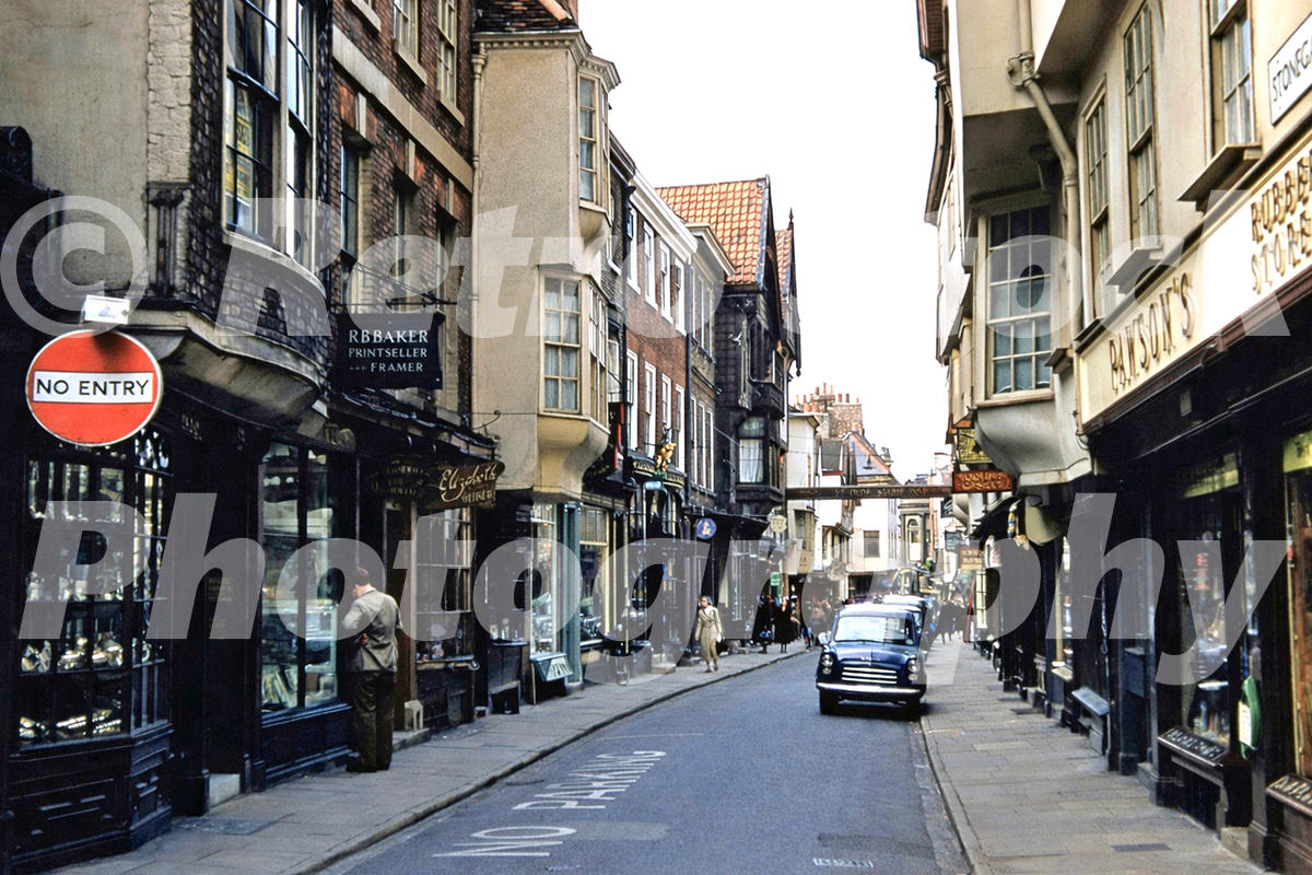 1950s view along Stonegate in York, with historic shopfronts, a No Entry sign, pedestrians and a parked black car.