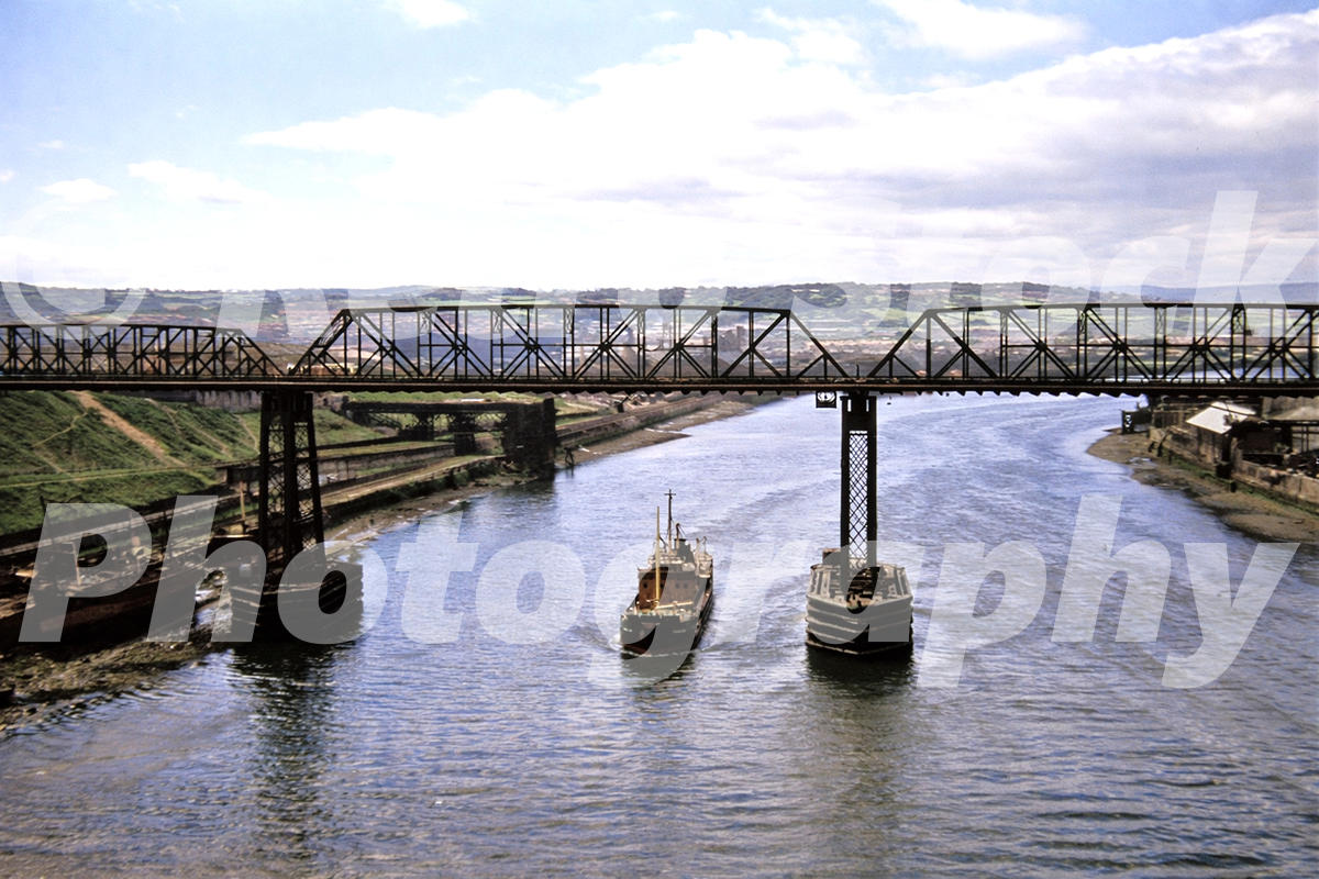 1950s colour photo of the old Redheugh Bridge over the River Tyne at Newcastle, with a small vessel passing beneath the steel spans.