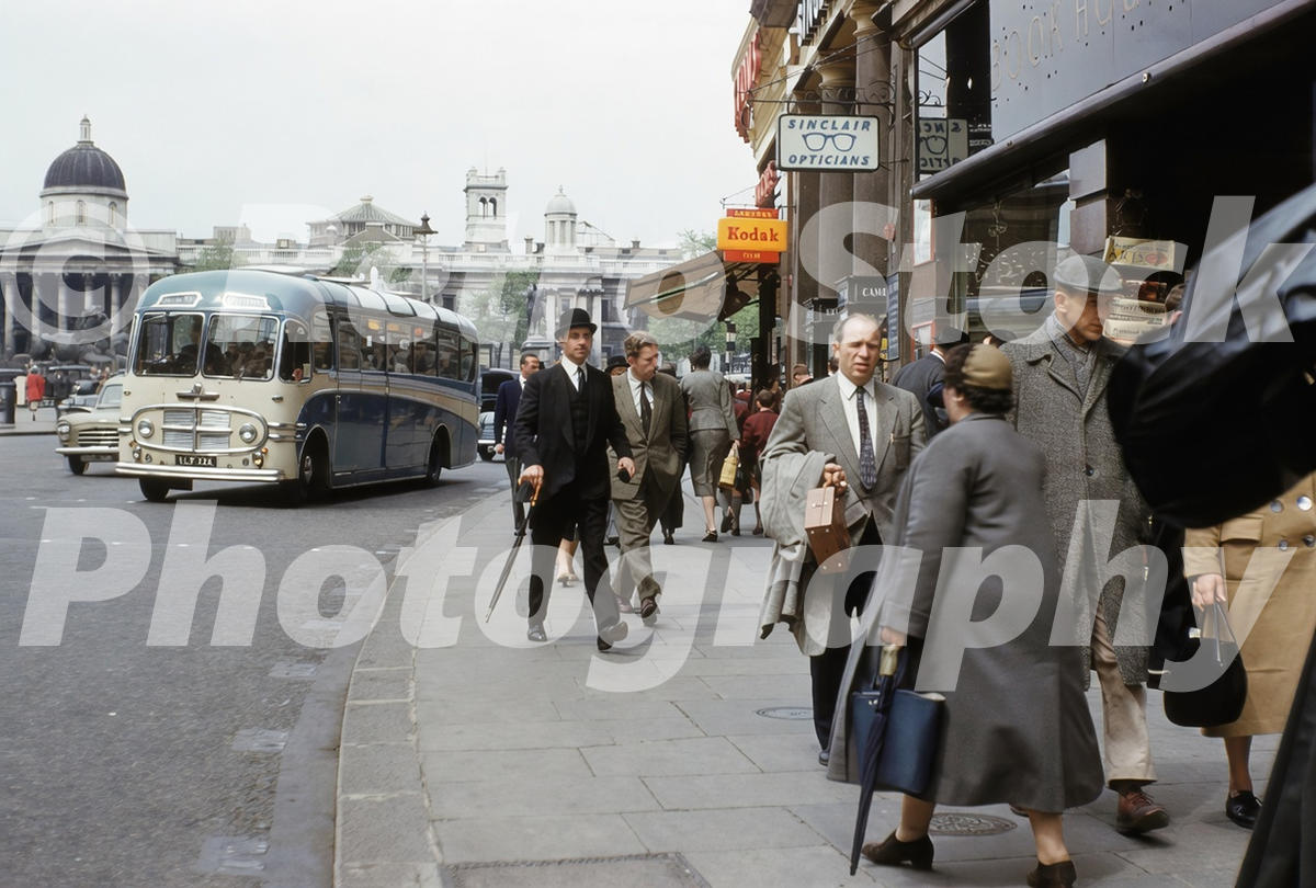 1950s colour photo of Trafalgar Square in London, with pedestrians, shop signs, a coach and the National Gallery in the background.