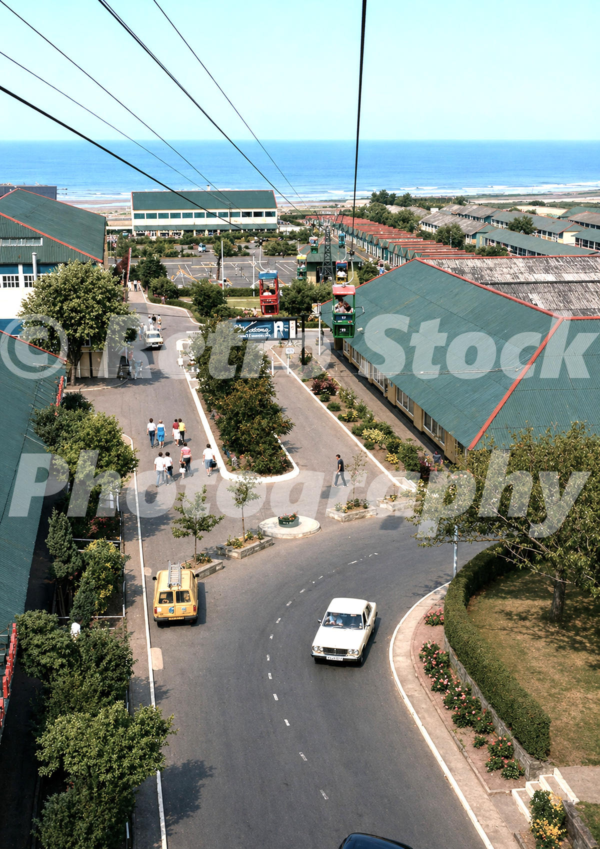 An aerial 1983 colour photograph taken from a chairlift overlooking a main thoroughfare within Butlins Ayr, showing holidaymakers walking between large green-roofed buildings and a white car driving below.