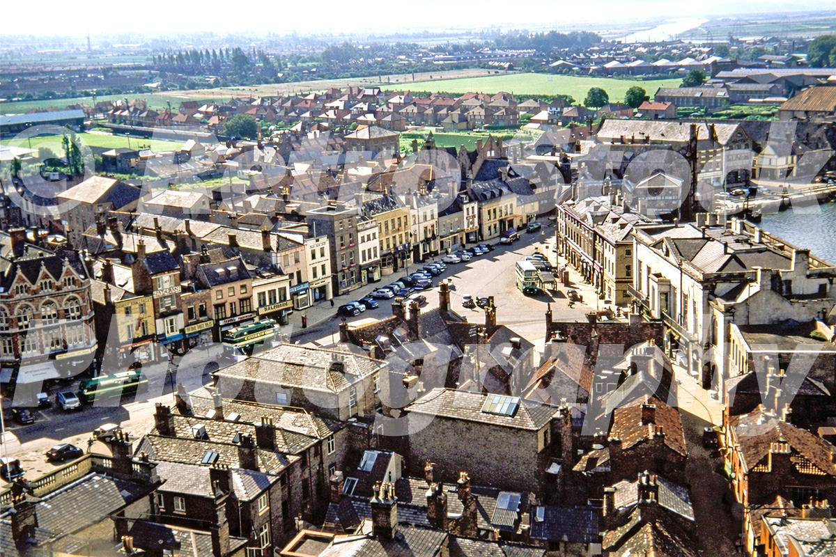 1950s colour photo of Boston Market Place in Lincolnshire, seen from St Botolph’s Church tower with buses, cars, shops and the River Witham beyond.