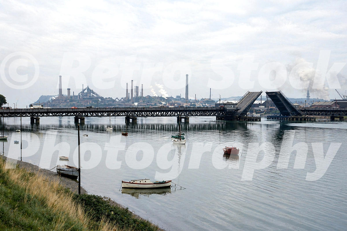1950s colour photo of Walney Bridge raised over the Walney Channel at Barrow-in-Furness, with small boats on the water and industrial chimneys in the background.