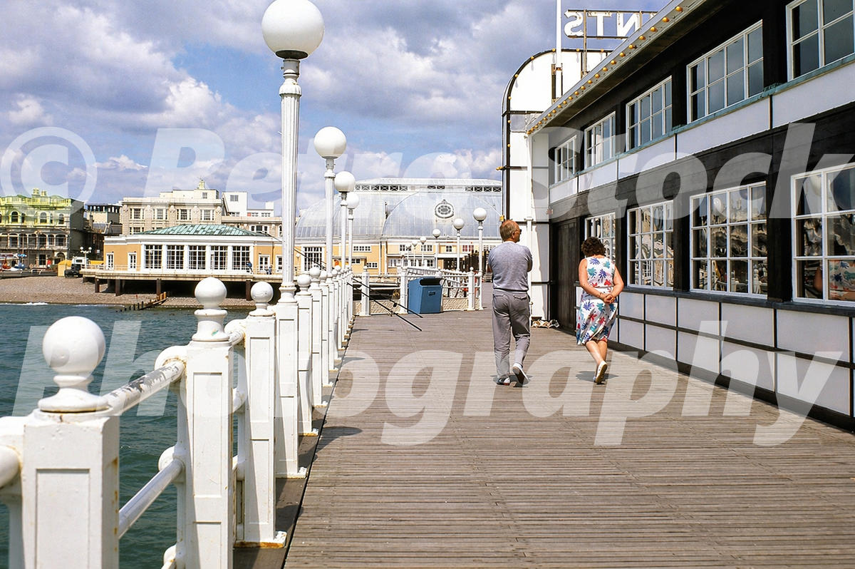 1980s colour photo of Worthing Pier, with people walking along the timber deck towards the Pavilion Theatre and seafront.