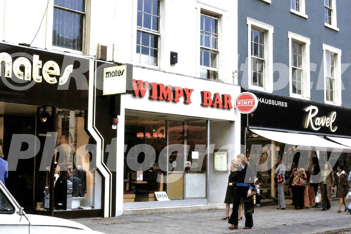 1970s view of a Wimpy Bar on King’s Road in London, with shopfronts, pedestrians, Mates and Ravel signs.