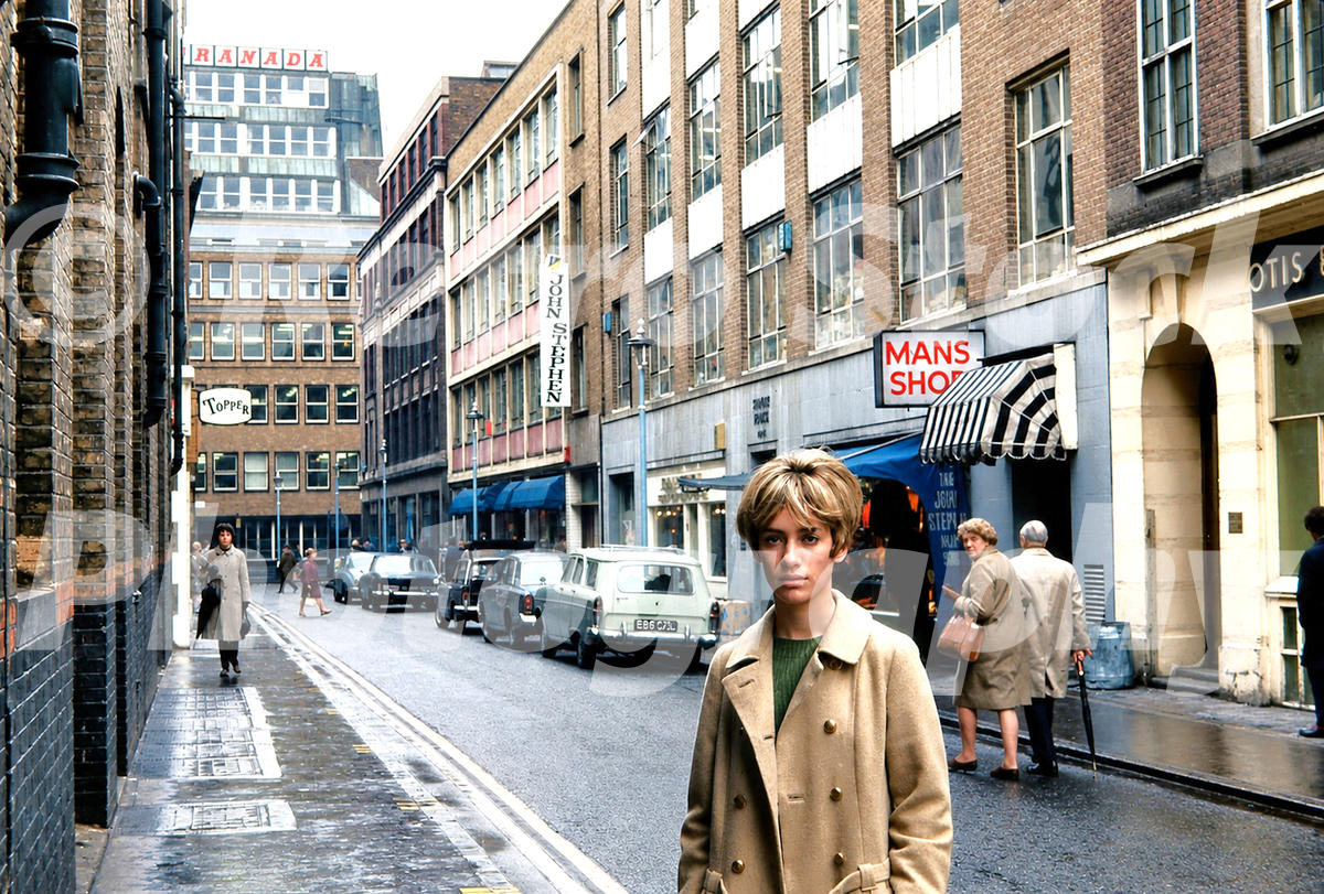 A 1960s colour photo of a young woman in a tan coat standing on Carnaby Street in 1966, with the John Stephen boutique in the background.