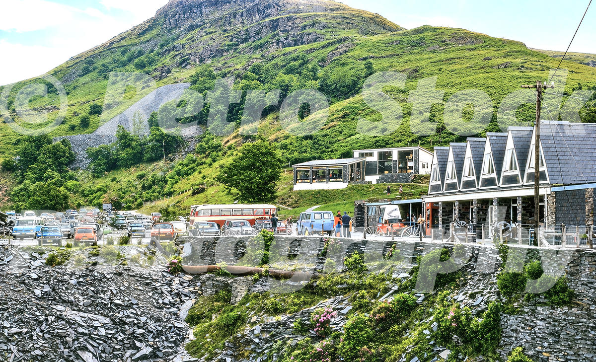 A 1977 view of Llechwedd Slate Caverns featuring the modern restaurant building with a saw-tooth roof, a red and cream coach, and slate tips on the mountainside.
