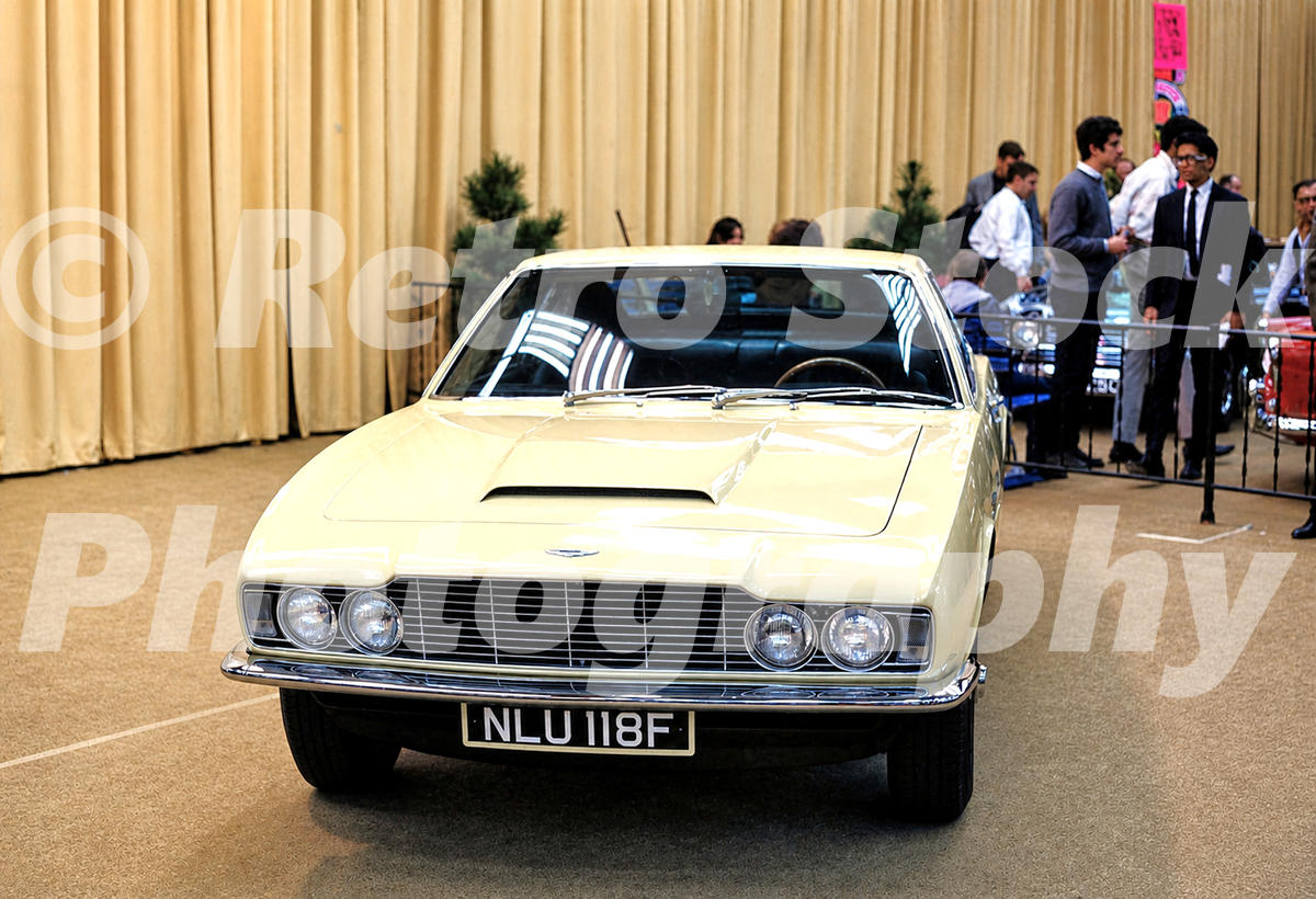 A 1960s colour photo of a Aston Martin DBS on display at the 1968 New York Auto Show, featuring its quad-headlight grille and British registration plate NLU 118F.