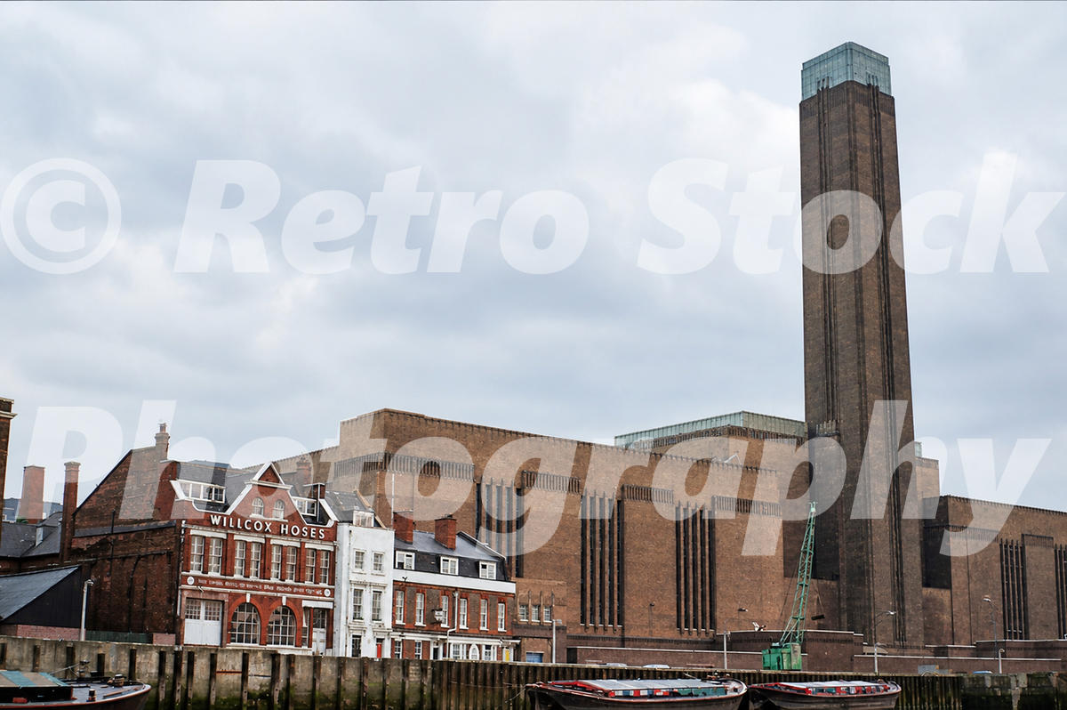 1960s view of Bankside Power Station beside the River Thames in London, with barges, the Willcox Hoses building and the tall power station chimney.