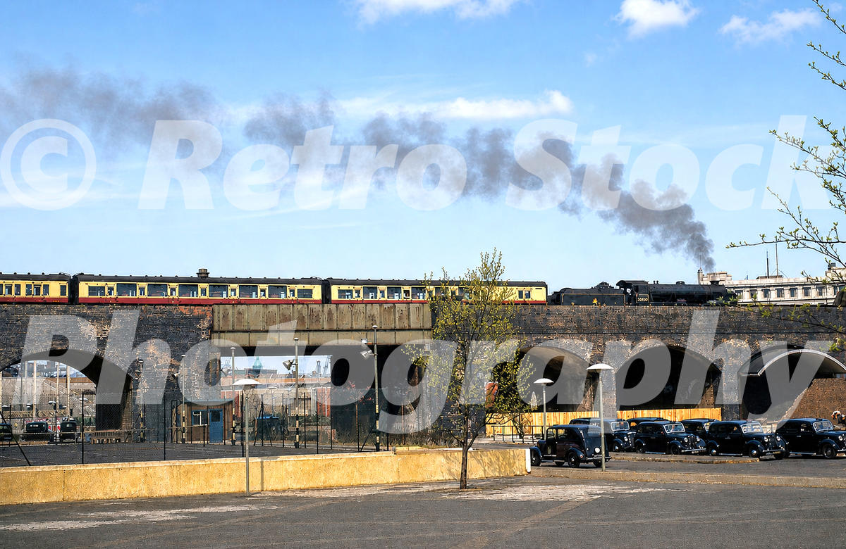 A 1950s colour photo of a steam locomotive and passenger train crossing a brick viaduct over Belvedere Road on the South Bank, London, with vintage black taxis parked below.
