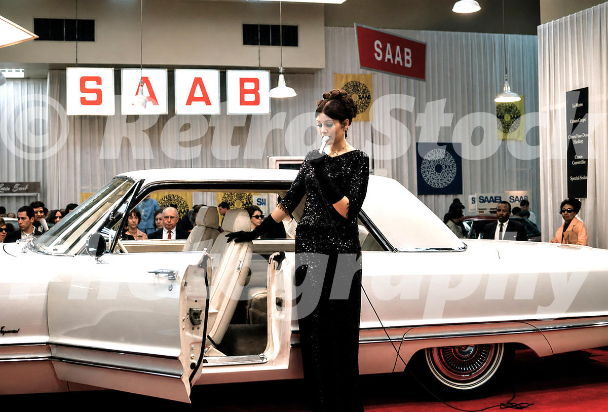 A 1960s color photo of a gold 1968 Cadillac Calais with a black vinyl roof on display at the New York Auto Show, with spectators and Volkswagen signage in the background.