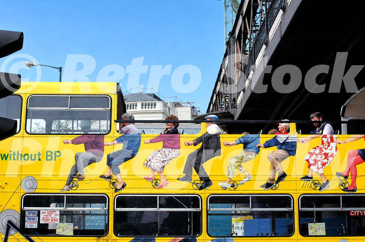1980s photo of a yellow Cityrama sightseeing bus in London, with an open top deck, painted cyclist-style figures along the side and an iron railway bridge above.