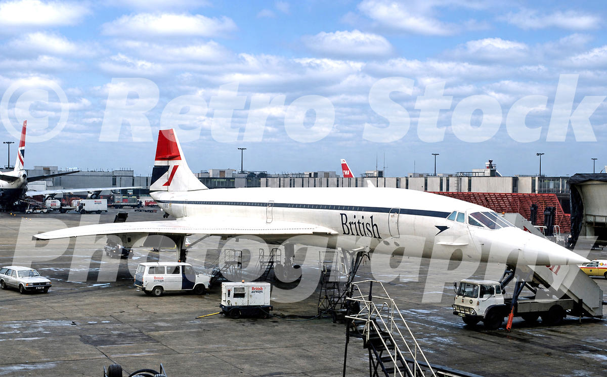 1980s view of a British Airways Concorde at Heathrow Airport, with ground service vehicles, passenger steps and terminal buildings behind.