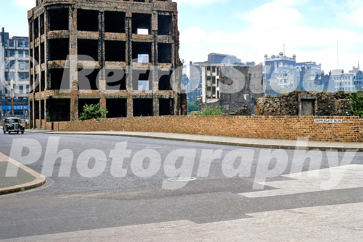 1950s colour view of bomb damage near Cripplegate Buildings in the City of London, with ruined walls, cleared plots, a brick boundary wall and a period car.