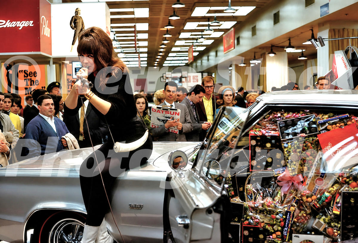A 1960s color photo of a promotional model in a black outfit sitting on a silver Dodge Charger filled with candy at the 1966 New York Auto Show.