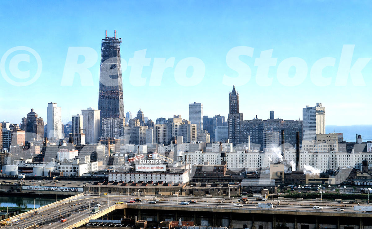 1960s color view of the downtown Chicago skyline, with the John Hancock Center under construction, expressway traffic, rooftop candy signs and Lake Michigan.
