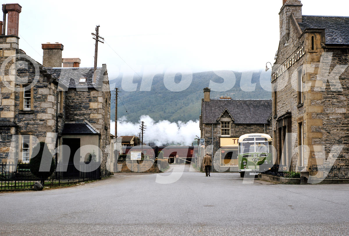 1950s view of Ford Road in Blair Atholl, Scotland, with the Atholl Arms Hotel, a green bus, stone buildings and train smoke by the level crossing.