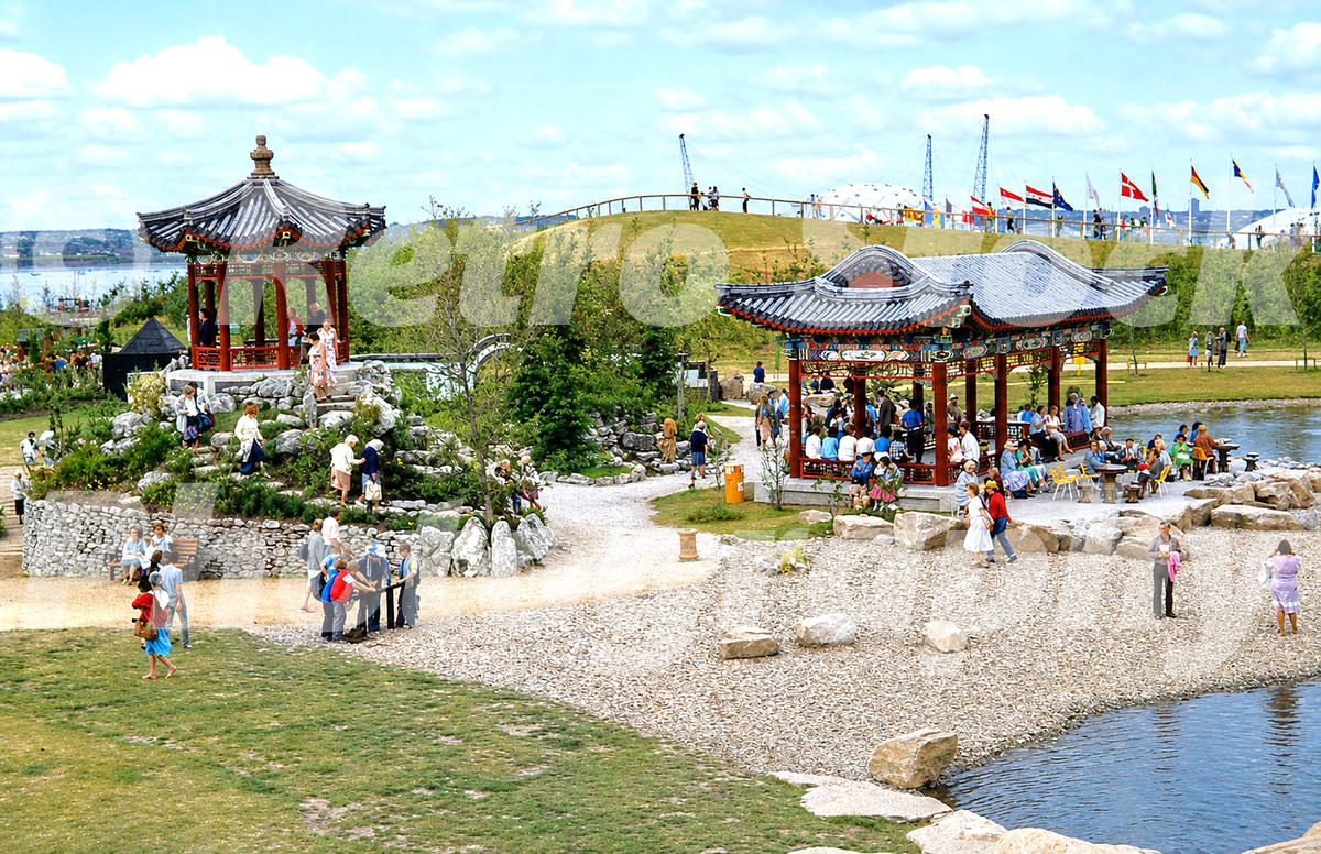 1980s view of the Japanese Garden at the Liverpool Garden Festival, with pavilions, rocks, water, visitors, flags and the River Mersey beyond.