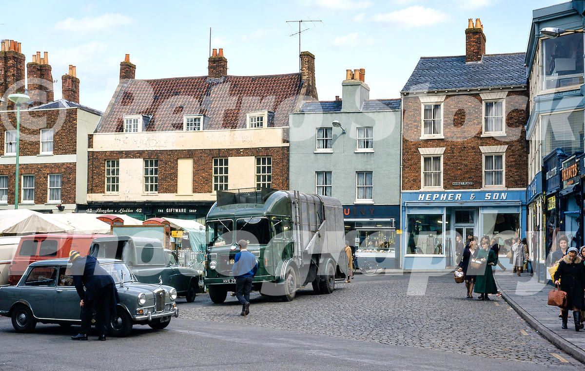 1970s view of Market Place in Wisbech, Cambridgeshire, with shops, market stalls, pedestrians, cobbles, a green lorry and period cars.