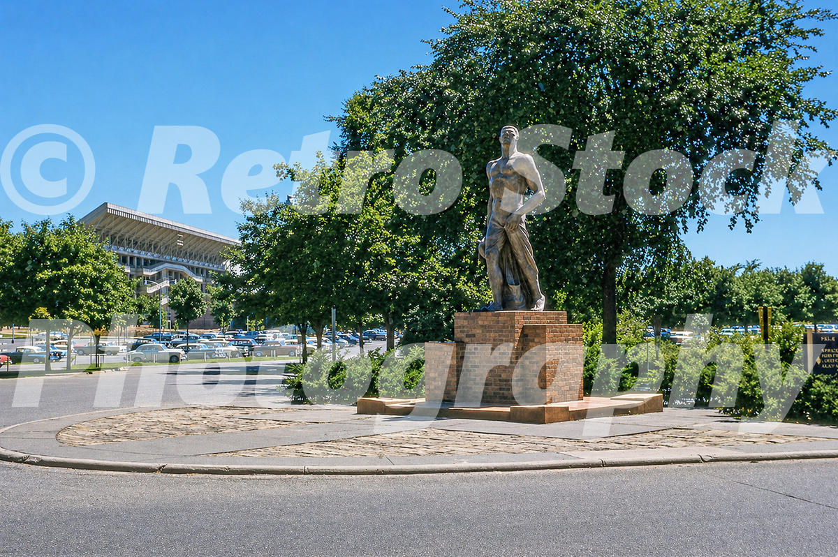 A 1950s color photo of the original terra cotta Spartan Statue on its brick pedestal at Michigan State University in East Lansing, with Spartan Stadium visible in the background.