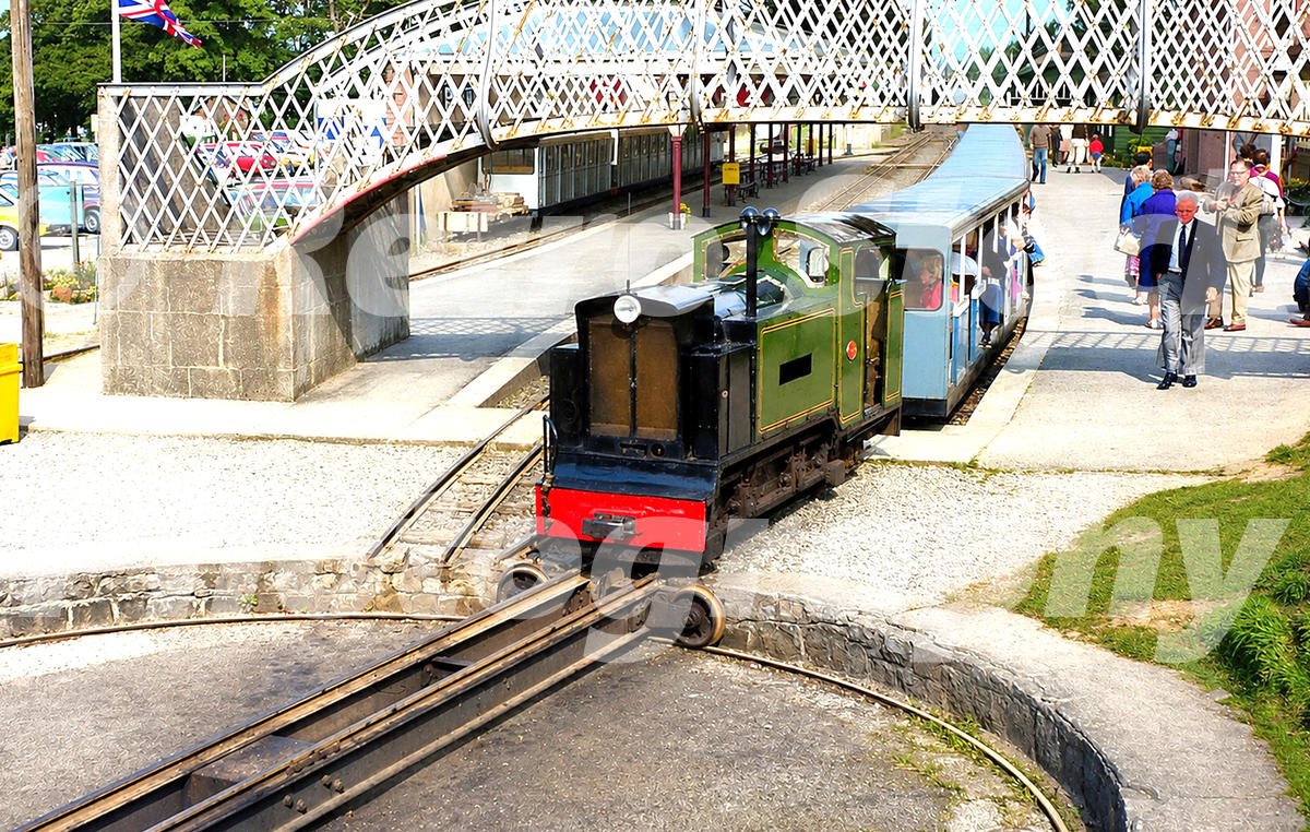 1970s view of a Muir-Hill Fordson locomotive at Ravenglass on the Ravenglass and Eskdale Railway, arriving with a passenger train beneath a footbridge.