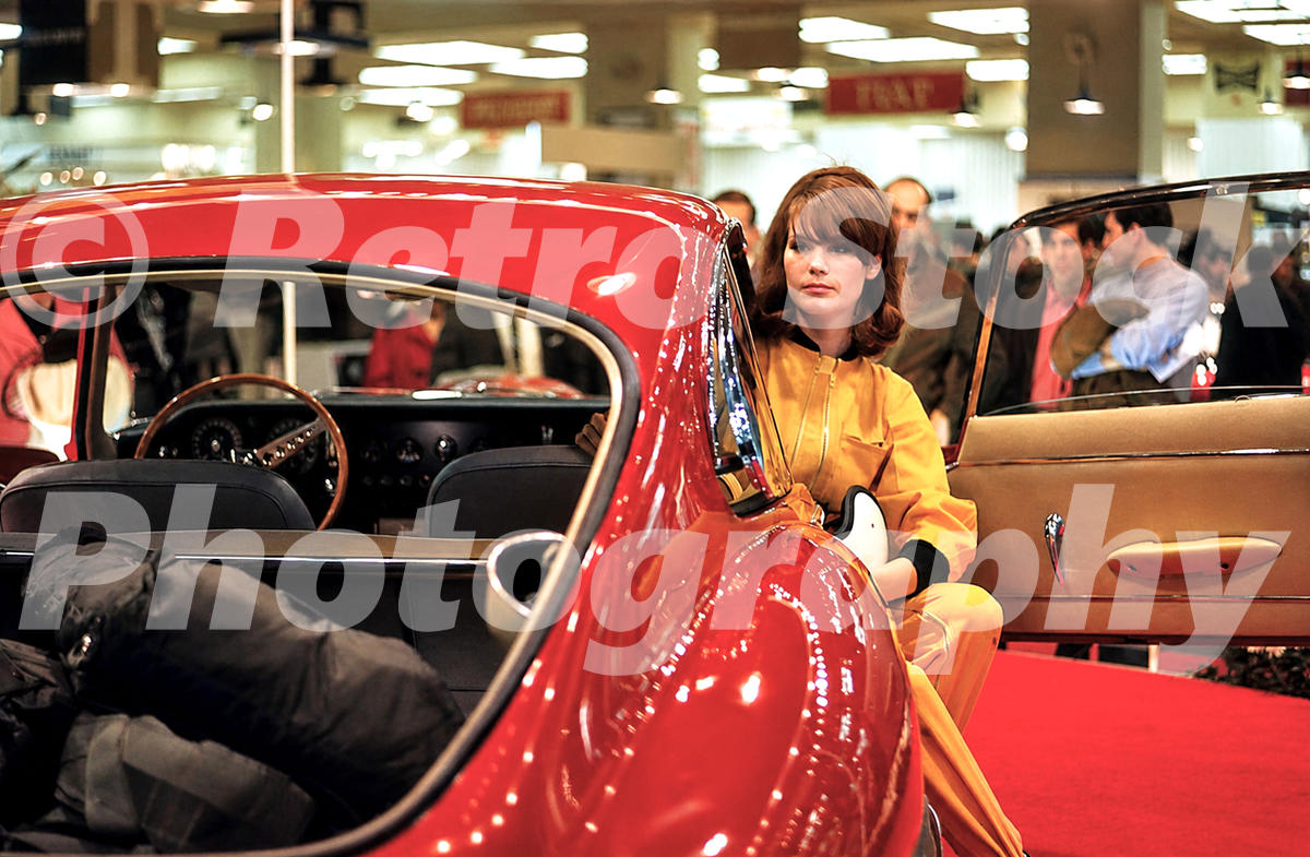 A 1960s colour photo of a red Jaguar E-Type Fixed Head Coupe with a model in a yellow jumpsuit standing by the open door at the 1966 New York Auto Show.