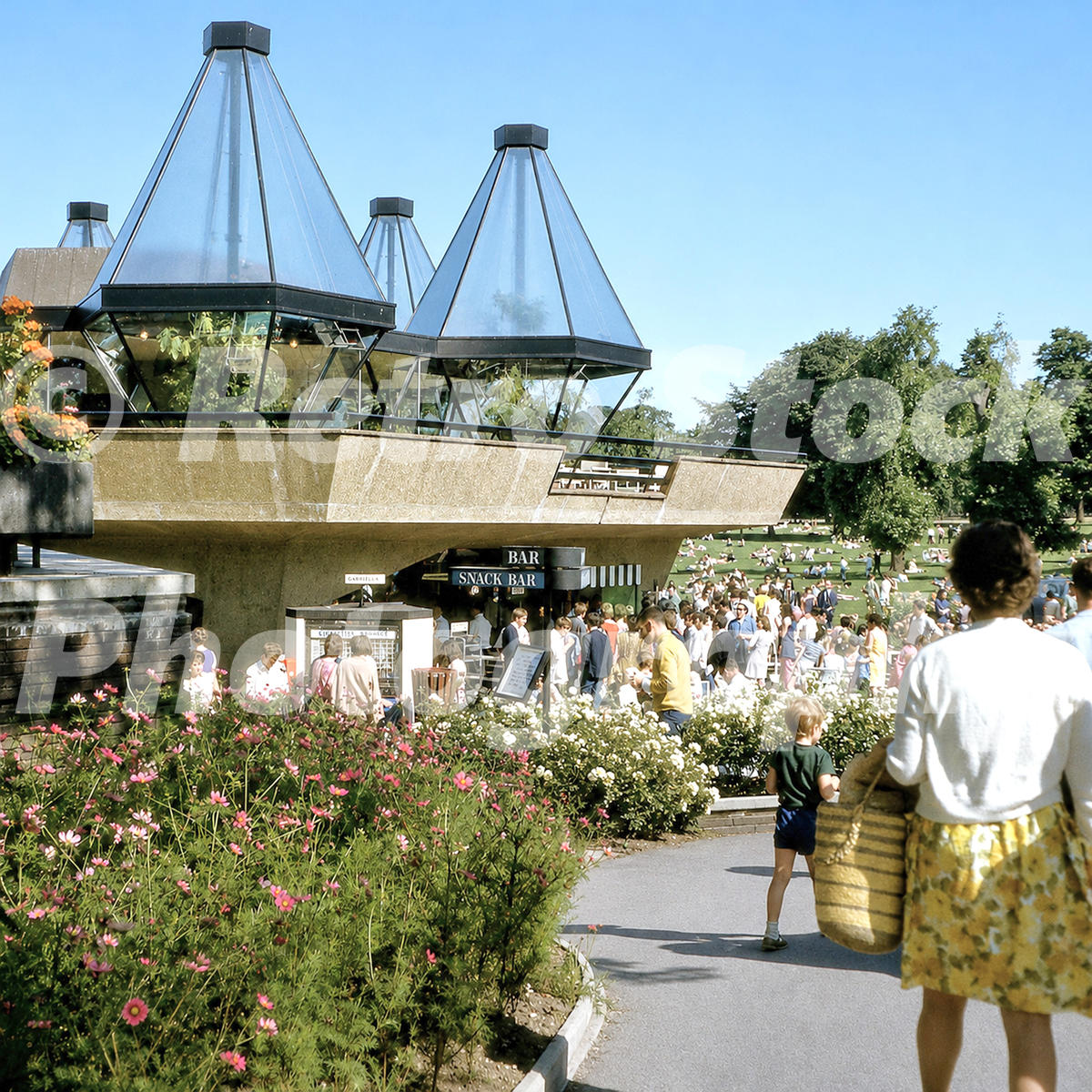 1960s view of the Serpentine Restaurant in Hyde Park, London, with modernist rooflights, flower beds, snack bar, visitors and people relaxing on the grass.