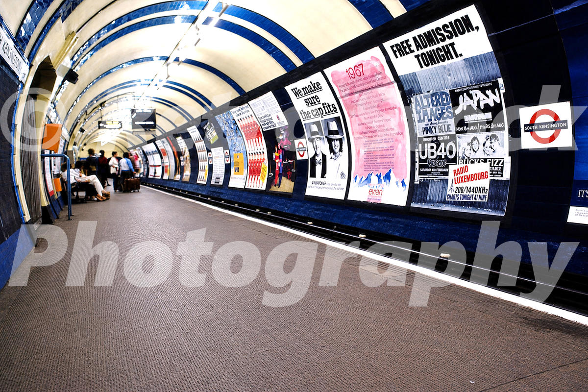 1980s view of South Kensington Underground station in London with a curved platform, cream and blue tiled tunnel roof and a long row of period advertising posters