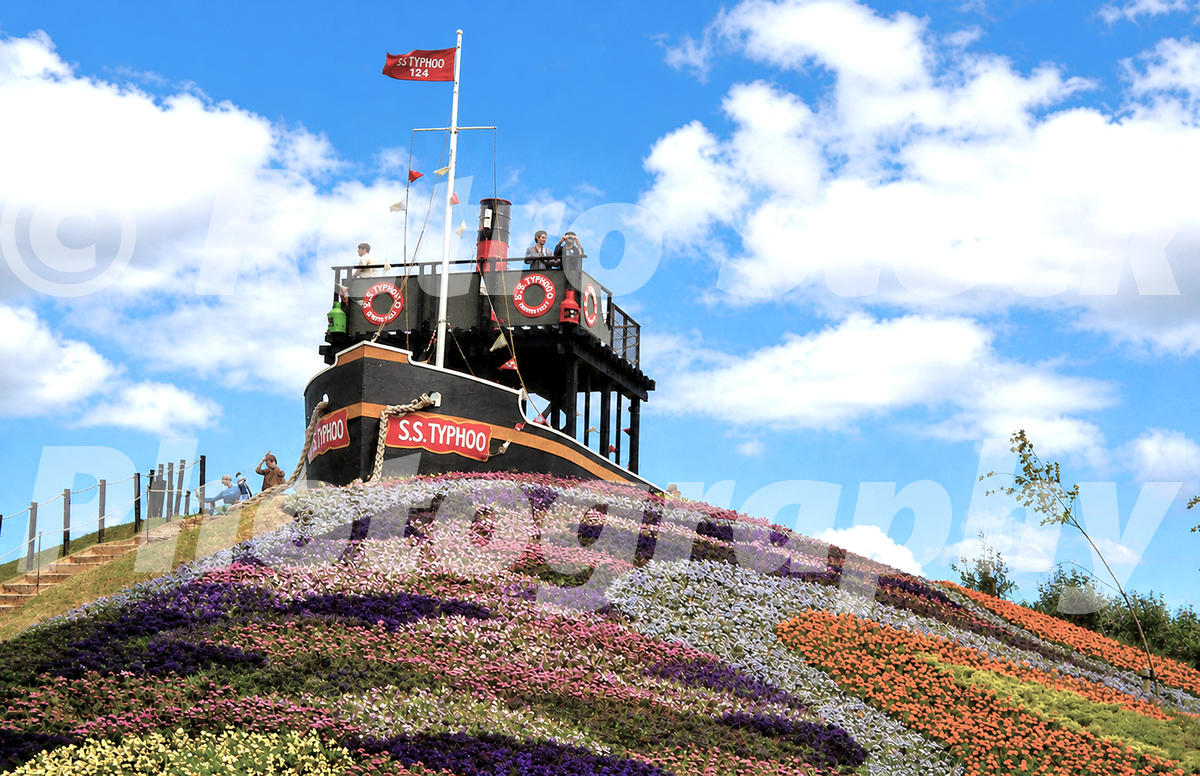 1980s view of the SS Typhoo display at the Liverpool Garden Festival, with a ship-shaped feature, colourful flower beds, flags and visitors.
